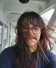 photo of Hannah Stohry smiling in the hallway of a boat with her long brown hair blowing in the wind. She is wearing brown rim glasses and a navy blue long sleeve top