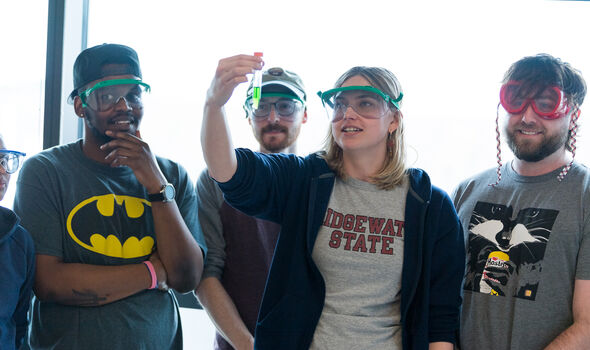 Four BSU students wearing safety goggles. One holds up a test tube with green liquid in it while the others look on.