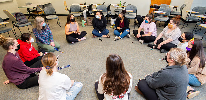 Professor Leslie Sattler and students sit in a circle on the floor of a carpeted classroom with one student speaking while everyone else looks on and listens