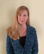 Maria Armour smiling with long dirty blonde hair parted on the side and wearing a black top under a blue, black, white and yellow print blazer