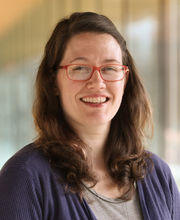 Jessica Birthisel smiling with long brown hair and wearing red rimmed glasses and a purple sweater over a gray top