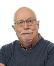 headshot photo of Dr. Walter Carroll, bald with a gray mustache and goatee wearing clear and light blue rimmed glasses and a blue v-neck sweater over a brown and blue button down shirt