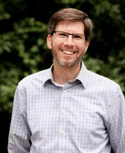 Ben Carson with short brown hair graying at the temples, brown and gray mustache and beard wearing glasses and a light blue button down shirt