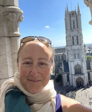 Dr. Jodi Cohen smiling with her brownish/red hair pulled back and sunglasses on top of her head. She is wearing a beige linen scarf and sleeveless purple top, standing on a balcony with a gothic church in the background