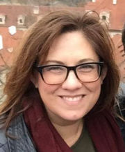 Amber Edwards smiling with long brown hair wearing black rim glasses, a black shirt and jacket and a maroon scarf