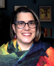 Dr. Kimberly Fox smiling with straight, medium length highlighted brown hair. She's wearing black rimmed glasses and a red, yellow, blue and green wrap