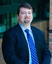 Professor Nicholas Ganson smiling with short brown hair, mustache and goatee wearing a navy blue blazer over a light blue button down shirt and blue striped tie