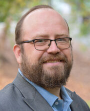 Dr. Feodor Gostjev smiling with short balding reddish brown hair, mustache and beard, wearing black rim glasses and a gray blazer over a light blue button up shirt