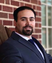 Dr. Kianoosh Hassani smiling with short dark brown hair, mustache and beard and wearing a navy blue suit jacket and tie with button down white shirt