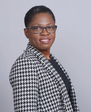 Dr. Myrlene Jean-Venant standing profile looking over her right shoulder smiling with short black and gray hair and black rimmed glasses. She is wearing a black, white and gray houndstooth blazer over a black top