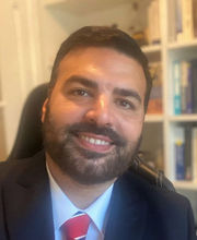 Dr. Chris H. Kennedy sitting in a desk chair with a built in book shelf in background. He is smiling, has short dark brown hair, beard and mustache and is wearing a navy blue suit jacket over a light blue button down shirt and red, white and blue tie