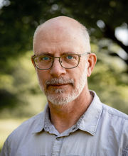 John Kucich balding with short gray hair, mustache and beard and wearing a light blue button up shirt with 2 front pockets