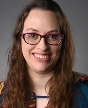 Rachael Linker smiling with brown rimmed glasses and long wavy brown hair and wearing a multicolored blouse