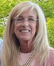 Dr. Kimberly Merrick smiling with long grey-blonde hair wearing rimless round glasses a pink top and pink pearls