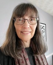 headshot photo of Dr. Erin O'Connor with long brown/grey hair and bangs smiling wearing brown rim glasses, a black top and purplish grey print scarf