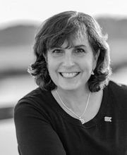 black and white photo of Lisa Rafferty smiling with shoulder wavy brown hair, leaning on her forearm and wearing a round neck dark colored three-quarter sleeve jersey