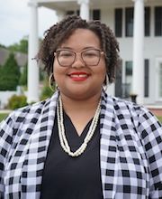 Dr. Sheena Rancher stands outside of the MRISJ office at 161 Summer street. She has short, curly hair and is wearing glasses with large earrings and a draped pearl necklace. Her blazer is black and white gingham