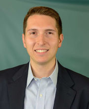 Philip Szlosek smiling with short brown hair and wearing a navy blue blazer over a blue and white pinstripe button up shirt