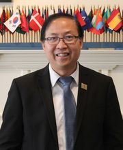 Dr. Wing-Kai To smiling with short graying black hair, wearing black rimmed glasses and a black suit jacket over a white button down shirt with a blue tie