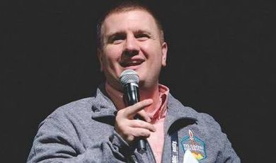 Mickey Howard stands in front of a black background holding a microphone