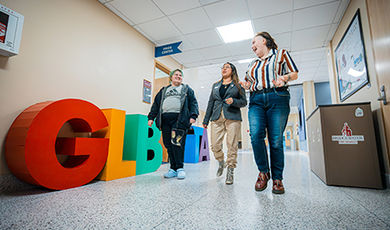 Three people walk down a hallway lined with large GLBTA letters