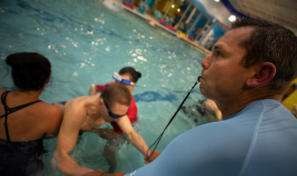 BSU student standing on side of a pool with a whistle in his mouth, bending over holding the hands of a male in the water who has 2 females on either side of him helping him move