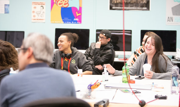 students in graphic design classroom smiling sitting around a table in the center of a room that has computers on desks around its perimeter
