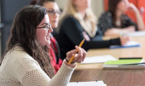 side view of a female BSU student with long dark brown hair and black wire rim glasses speaking, holding a pencil, sitting at a table with 3 other students