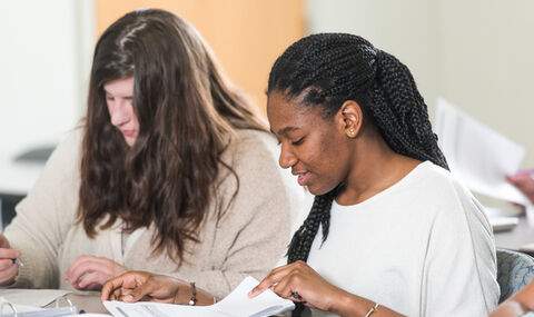 2 female students sitting at a classroom desk reading papers and writing