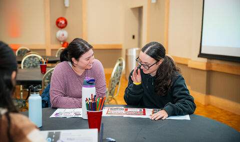 Two women sit at a table looking at and talking about materials on the table with BSU balloons in background