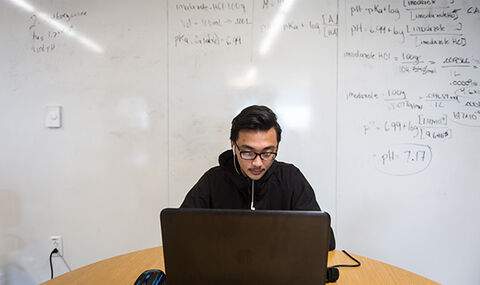 a BSU chemistry student working at a laptop in front of a large white board with columns of Chemistry notes titled Have, Need and Know