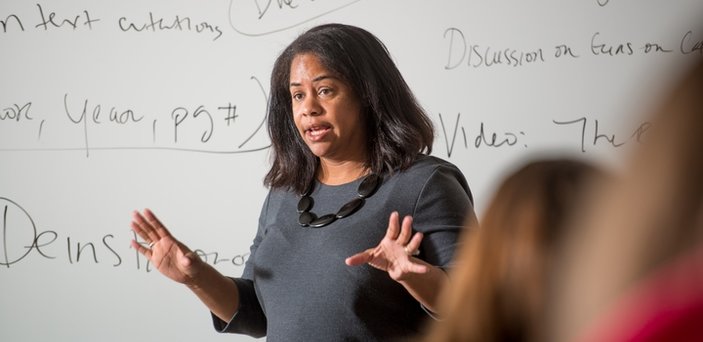 A professor teaching class in front of a white board with notes including "Discussion on guns" and "text citations."