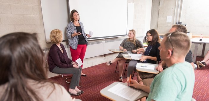 A professor speaking in front of a screen surrounded by a circle of students in desks listening