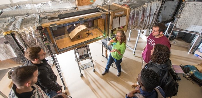 A professor with a group of students in front of the wind tunnel