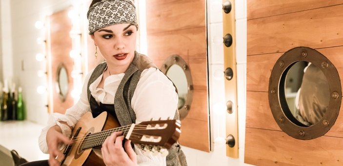 An actress playing a pirate who is playing guitar in front of portholes 