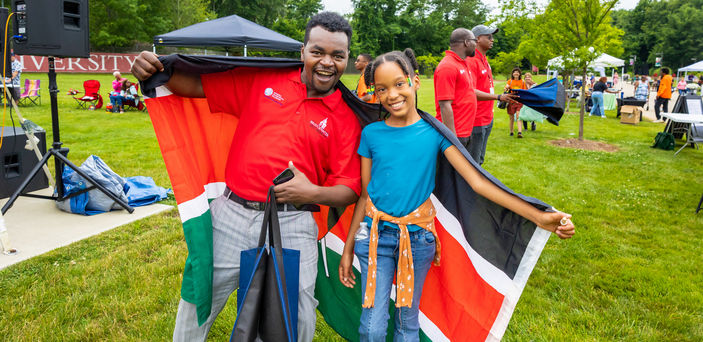 A Mandela fellow and child pose for a photo with a flag draped behind them.