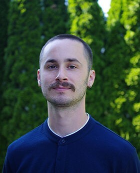 Headshot photo of Professor Eric Aldieri with very short brown hair and mustache wearing navy blue jersey with white trim collar in front of tall green bushes