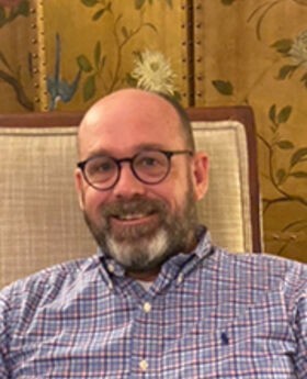 Dr. Jordon Barkalow sitting in a chair smiling with balding light brown hair, a beard and mustache wearing brown rimmed glasses and a blue checked button down shirt
